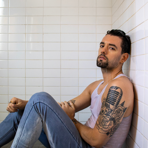 A muscular white man leans against the wall on the floor of a white-tiled shower. He wears a white tank top and blue jeans and has an elaborate tattoo of a microphone on his arm. He has black hair slicked back with water and a manicured beard.