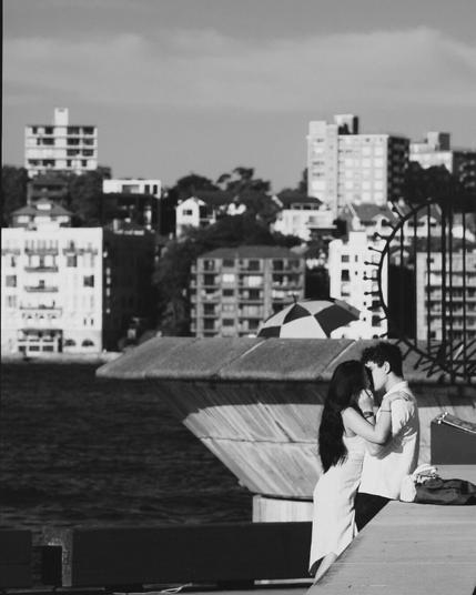 This black-and-white image shows a couple embracing and kissing near a waterfront. In the background, you can see buildings and structures across the water. The scene appears to be urban, with a mix of residential and possibly commercial buildings. The setting suggests a moment of intimacy in a busy city environment.