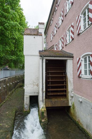 Auf dem Foto ist ein altes hölzernenes Mühlrad abgebildet, das unter einem Gebäude in einen Fluss ragt.