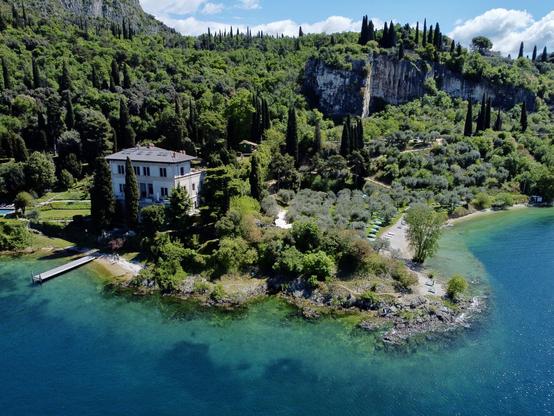 Ein felsiger Strandabschnitt mit Bergen im Hintergrund und starkem Bewuchs. Das Wasser in Ufernähe zeigt verschiedene Blau- und Türkistöne. Eine kleine Villa steht nahe am Strand, ein kleiner Steg erstreckt sich von der Villa ins Wasser
