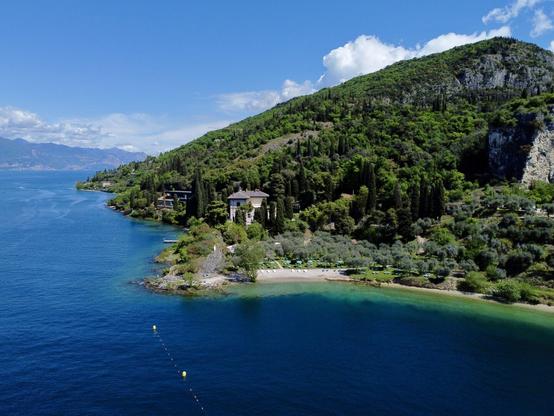 Eine kleine felsige Landzunge ragt in den Gardasee. Im Hintergrund ist ein stark bewachsener Berg zu sehen. Etwas versteckt von Vegetation steht eine kleine Villa in Ufernähe.