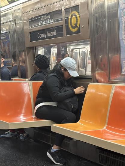 NYC subway Q train, sign says Coney Island bound. A young woman with long dark hair and a grey ball cap which hides her face sits beneath, in a black track suit and black sneakers.