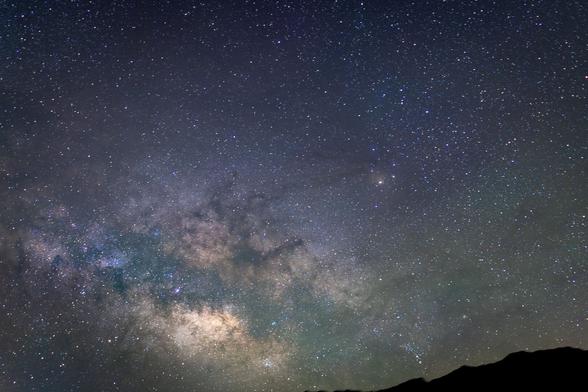 Starry night sky with pale band of Milky Way across bottom left corner with silhouette of hill on bottom right