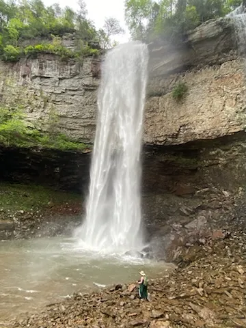 Photo of a very tall waterfall with heavy,  high pressure flow. It falls over a rock ledge into a stoney pool. A small elf in a green dress and sun hat stands at the edge of the pool.