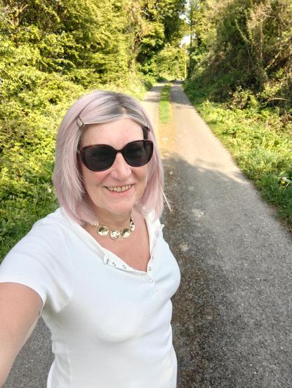 A pink haired woman standing in a country lane with grass growing up the middle of it.
I'm wearing a white henley neck top, gold disc necklace and sunglasses.