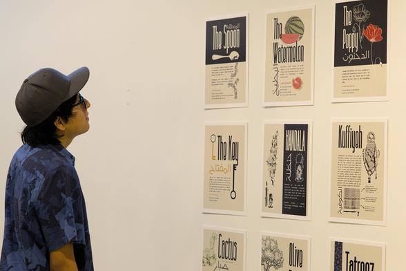 Person in a dark blue patterned shirt and black cap looking at a wall. The wall features 6 different posters representing different aspects of Palestine and Palestinian resistance: the spoon, the watermelon, the poppy, the key, Handala, kuffiyeh, cactus, olive tree, and tatreez