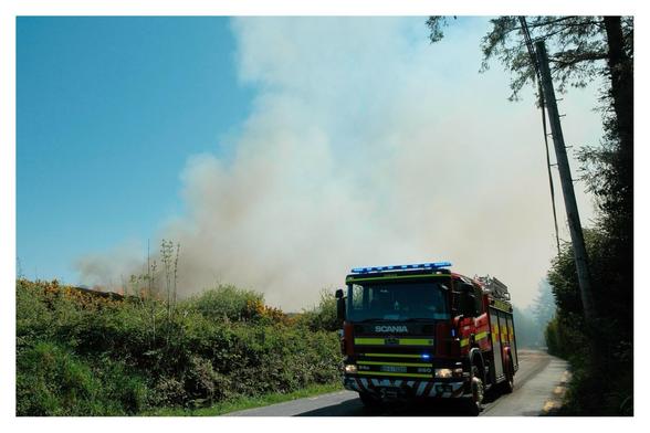 Fire truck on the road next to a fire & smoke rising into the blue sky.