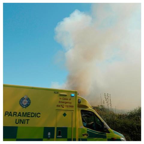 Ambulance on the road next to a fire & smoke rising into the blue sky.