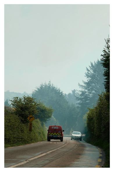 Fire support vehicle heading down the road into the hazy smoke filled air. A car approaches in the opposite direction