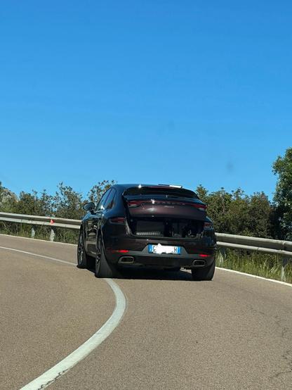 Porsche Macan with half-open trunk on a mountain road. In the trunk are several suitcases and smaller bags just waiting to fall out.