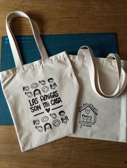 Dos bolsas de tela cruda sobre una mesa de madera. Una tiene la frase “LAS AMIGAS SON MI CASA” acompañada de dibujos de rostros sonrientes; la otra muestra un dibujo de una casa con el texto “con las amigas y en Conce”.