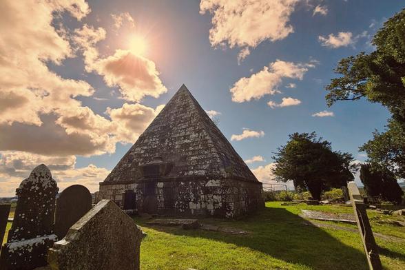 A stone pyramid-shaped mausoleum stands in a grassy graveyard under a partly cloudy sky with the bright sun visible. Several weathered gravestones are visible in the foreground