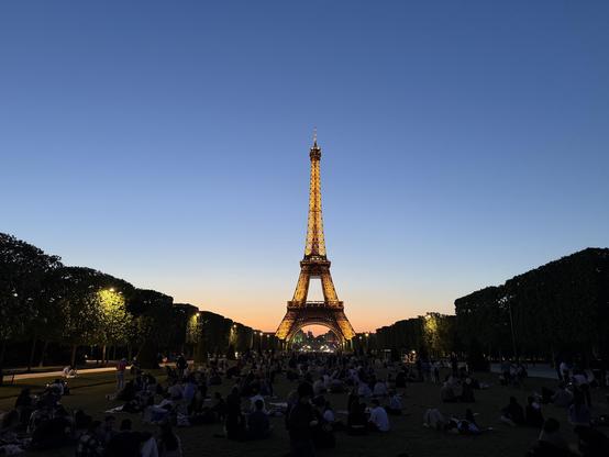 La Tour Eiffel après le couché de soleil. La tour est illuminé, mais le ciel est encore bleu derrière et part sur un dégradé orange en bas. En dessous, dans l’obscurité, on voit le Champ de Mars avec beaucoup de monde qui admire le monument. On voit des arbres sur les deux côtés qui donnent un effet de perspective avec un point de fuite au pied de la tour.