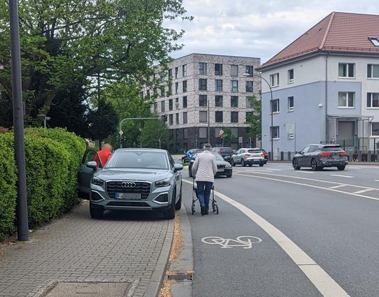 Auf einem Fußweg wurde ein SUV so abgestellt, dass der Gehweg komplett blockiert ist. Aus dem Auto steigt eine Person auf der Beifahrerseite aus, während eine ältere Person mit Rollator auf der Fahrbahn neben dem Auto her läuft - auf einem Radfahrstreifen in Gegenrichtung.