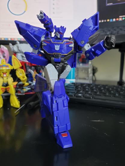 A photo of Transformers Earthspark Deluxe-class Soundwave posed walking across a desk. There are computer monitors, a keyboard, and other Transformers toys in the background.