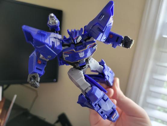 A photo of Transformers Earthspark Deluxe-class Soundwave in a flying pose, being held up by a human hand. There are a computer monitor and the edge of a windowframe in the background.