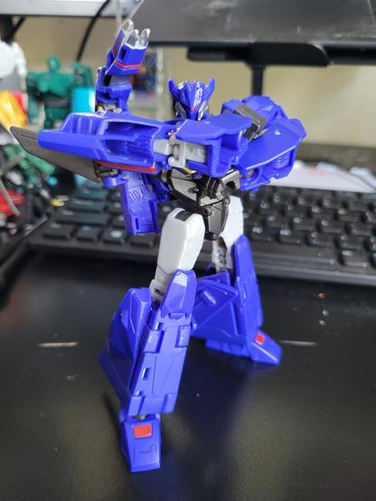 A photo of Transformers Earthspark Deluxe-class Soundwave posed standing with arms crossed, on a desk. There are a computer keyboard and other Transformers toys in the background.