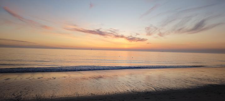 Bild eines Sonnenuntergangs am Strand. 
Der Sand ist schon halb im Dunkeln versunken, in der Brandung spiegelt sich das letzte orangfarbene Licht. Der Himmel hat alle Schattierungen von hellem blaugrau über gelb, orange und rot-Töne, die sich im Meer spiegeln. Spärliche dünne graue Wolken davor