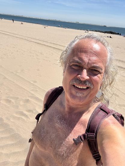 Selfie of me on the beach; long white hair, mustache and jazz patch, hairy chest. All my stuff is in my backpack, the sand and ocean are seen over my shoulder.