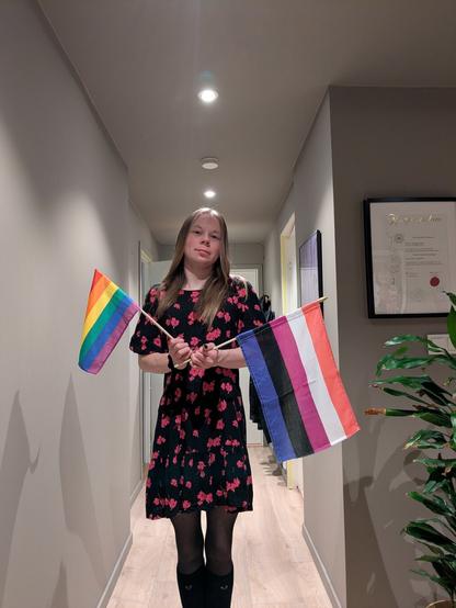 A woman stands in her apartment.

She is holding a LGBTQ+ flag and a genderfluid flag. 

In the foreground, the woman stands in a hallway with an indoor plant visible to the right and a certificate on the wall behind.