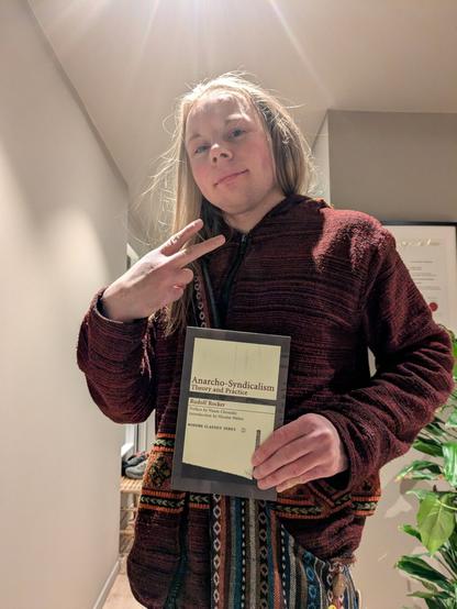 The image presents a young man standing within an apartment.

He is holding a book titled "Anarcho-Syndicalism: Theory and Practice" by Rudolf Rocker.

A plant and a framed certificate are visible in the background.