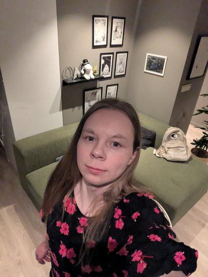 A woman in their mid-twenties, poses for a selfie in her apartment.

Behind them, a shelf displays various figurines and framed artwork.

In the foreground, a sofa and a table can be seen in her living room.