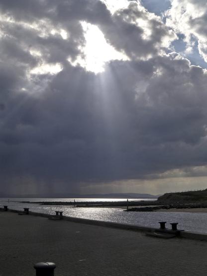 Dramatic dark clouds with a break in purest white from which radiate many rays. Further out at sea, veils of rain are suspended from those same clouds. The sea surface is reflecting the brilliant sunlight. The pier in the foreground, with its old cast-iron bollards running allong the waterline along a diagonal from low on the right hand side , is in stark contrast. The further side of the harbour mouth a sandy with screes of small black rocks.The backlight turns it almost into an outline. The silhouette of a mountainous island is visible above the horizon.
