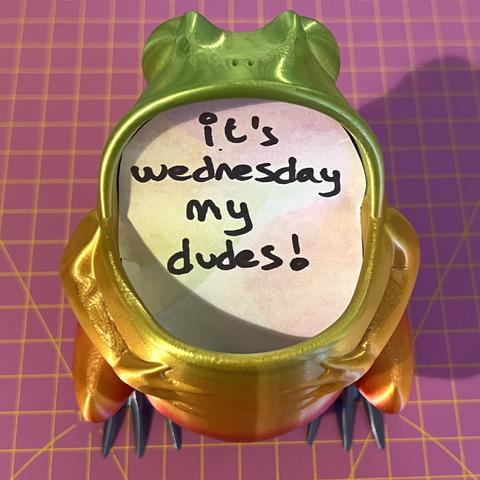 A 3D printed frog with a wide open mouth. A piece of paper is inside with "it's Wednesday my dudes" written on.