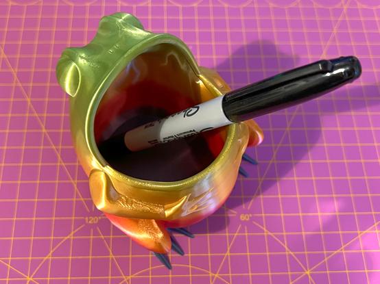A 3D printed pencil pot in the shape of a frog with a large open mouth. It has been printed in a rainbow silk filament. A black Sharpie pen is inside the pot.