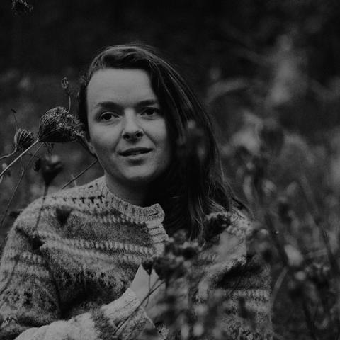 A lack and white phot of a non-binary person with long dark hair, a nose ring, and a striped sweater standing in tall grass.