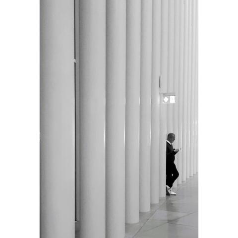 Black and white candid photo of man dressed in black, partly hidden among the white pillars of the Philharmonie building in Luxembourg.