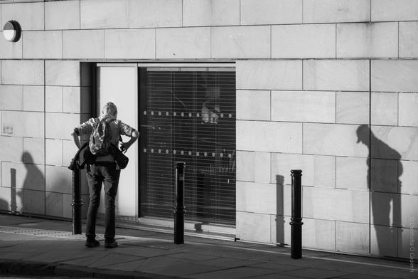A black and white street photograph taken in Edinburgh, Scotland, showing a person with a backpack standing with hands on hips, facing a large window set in a tiled wall; their shadow and the shadows of bollards are cast sharply on the wall, with another prominent shadow of the person to the right; a round wall light is visible in the top left.