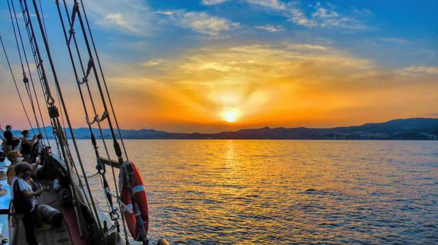Aquesta imatge captura la bellesa serena d'una posta de sol des del costat d'un vaixell de vela. Els colors càlids de la posta del sol pinten el cel amb tons daurats i taronges, contrastant bellament amb el blau profund del mar. El sol, gairebé amagat darrere d'una línia de muntanyes llunyanes, projecta raigs de llum que s'estenen pel cel.

L'escena transmet una sensació de pau i tranquil·litat. Els passatgers del vaixell, amb prou feines visibles, semblen estar gaudint de l'espectacle. La presència del socorrista en primer pla afegeix un toc subtil de seguretat, mentre que les cordes i l'aparell del vaixell suggereixen aventures i viatges.
