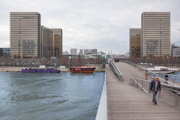 A man walks on a modern pedestrian bridge over a river, with boats docked along the shore and four glass library towers in the background under a cloudy sky.