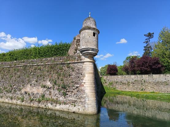 Photographie numérique couleur en format paysage pour les 2 premières photos, en format portrait pour les 2 suivantes.
Des douves remplies d'eau au premier plan, dominées par une fortification en pierre et en pointe surmontée d'une échauguette.
Une autre partie de rempart, des  arbres au-delà et un ciel bleu et lumineux avec quelques nuages blancs.