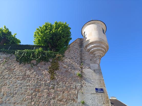 Une échauguette en plan plus serré vue en contre-plongée avec le mur en pierre claire en dessous, des arbres et du lierre en haut du mur. Il y a une plaque de rue et un ciel entièrement bleu.