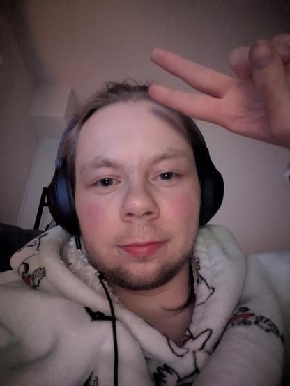 The image captures an adult person indoors. In the foreground, the person is wearing headphones and a patterned hoodie. A hanger is vaguely visible behind the person.