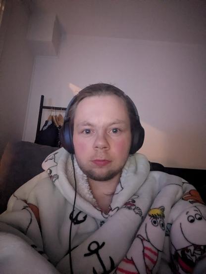The image captures an adult person indoors. In the foreground, the person is wearing headphones and a patterned hoodie. A hanger is vaguely visible behind the person.