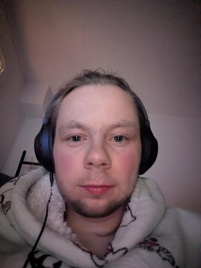The image captures an adult person indoors. In the foreground, the person is wearing headphones and a patterned hoodie. A hanger is vaguely visible behind the person.