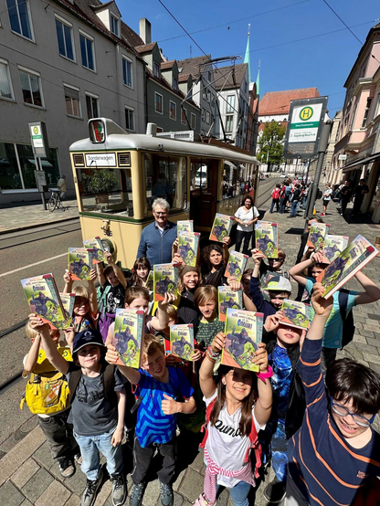 ine Gruppe fröhlicher Grundschulkinder steht bei sonnigem Wetter vor einer historischen Straßenbahn in der Augsburger Innenstadt. Jedes Kind hält stolz ein Exemplar eines Kinderbuchs in die Höhe. Im Hintergrund sieht man alte Stadthäuser, ein Haltestellenschild und Erwachsene, darunter der Vorleser Volker Nauerz, der Chef der Augsburger Stadtwerke. Die Stimmung ist lebendig und begeistert.