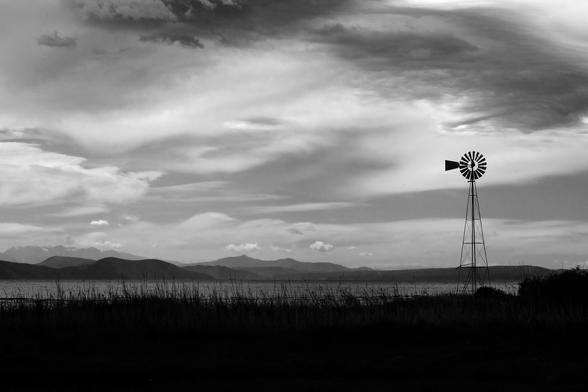 Black-and-white photo of a steel-framework water-pumping windmill on a sea coast. In the distance are several mountains, some with snow-covered peaks.