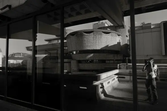 Noir et blanc. Reflet d'immeubles dans une grande baie vitrée. Le reflet du photographe est visible à droite de l'image.
