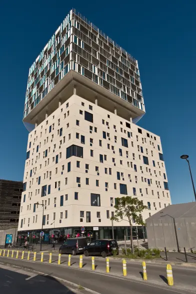 Photo verticale d'une tour composée de deux blocs distincts. Le second repose sur des pilotis sur le toit du premier. Les deux ont un aspect très différent.
