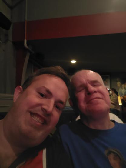 I think this was inside the Brattle?

Be My AI: Two men are sitting close together, smiling for a selfie. The man on the left has short brown hair and is wearing a dark shirt with red and white stripes on the shoulder. The man on the right has short gray hair and is wearing a blue shirt with a graphic of a person's face on it. His eyes are closed, and he has a content expression. The background is dimly lit, with a ceiling light visible above them and some framed pictures on the wall. I'm the guy on the right. Speaking of "lovestruck". LOL!
