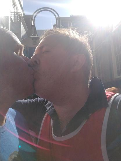 Be My AI: Two men are kissing each other. The man on the right is wearing a red and white top, while the man on the left is wearing a blue shirt. The sun is shining brightly in the background, creating a lens flare effect across the image. The setting appears to be outdoors, possibly in an urban area, with buildings and a street lamp visible behind them.