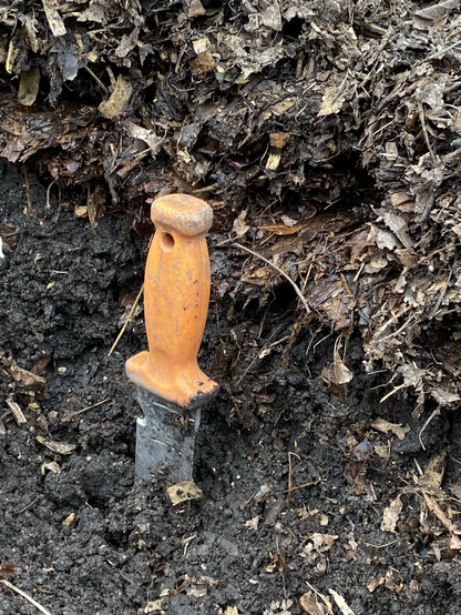 A side view of two layers in the compost pile. The top is a thick layer of brown and tan leaves.  The bottom is near black soil. The hori hori is vertical, blade silver, handle orange.