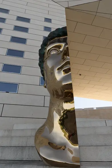 Sculpture d'une moitié de buste géant. La ligne de coupure s'aligne parfaitement avec le bord du batiment. Le matériau reflète l'environnement.