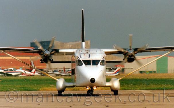 Head-on view of a white, boxy, high-winged, twin propellor-engined airliner parked facing the camera.
Sponsons sticking out from the bottom of the fuselage contain the main wheels, and are connected to the undersides of the wings by long thin struts.
Spinning propellors on the engines are blurred, not quite becoming smears.
In the background, a varied array of light aircraft are parked facing in different directions, with a couple of brown and grey buildings at the back, under grey skies.