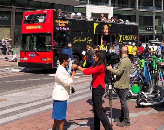 A person speaks with a TV crew in front of a human-protected bike lane. A double-decker bus is stopped next to the line of people acting as a bike lane barrier.