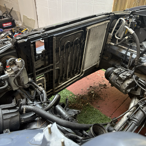 A view into the mostly empty engine compartment of my 2003 BMW 5-series station wagon, where the A/C condenser and transmission and power steering coolers have been installed into a new black plastic carrier just behind the radiator support across the front of the car.